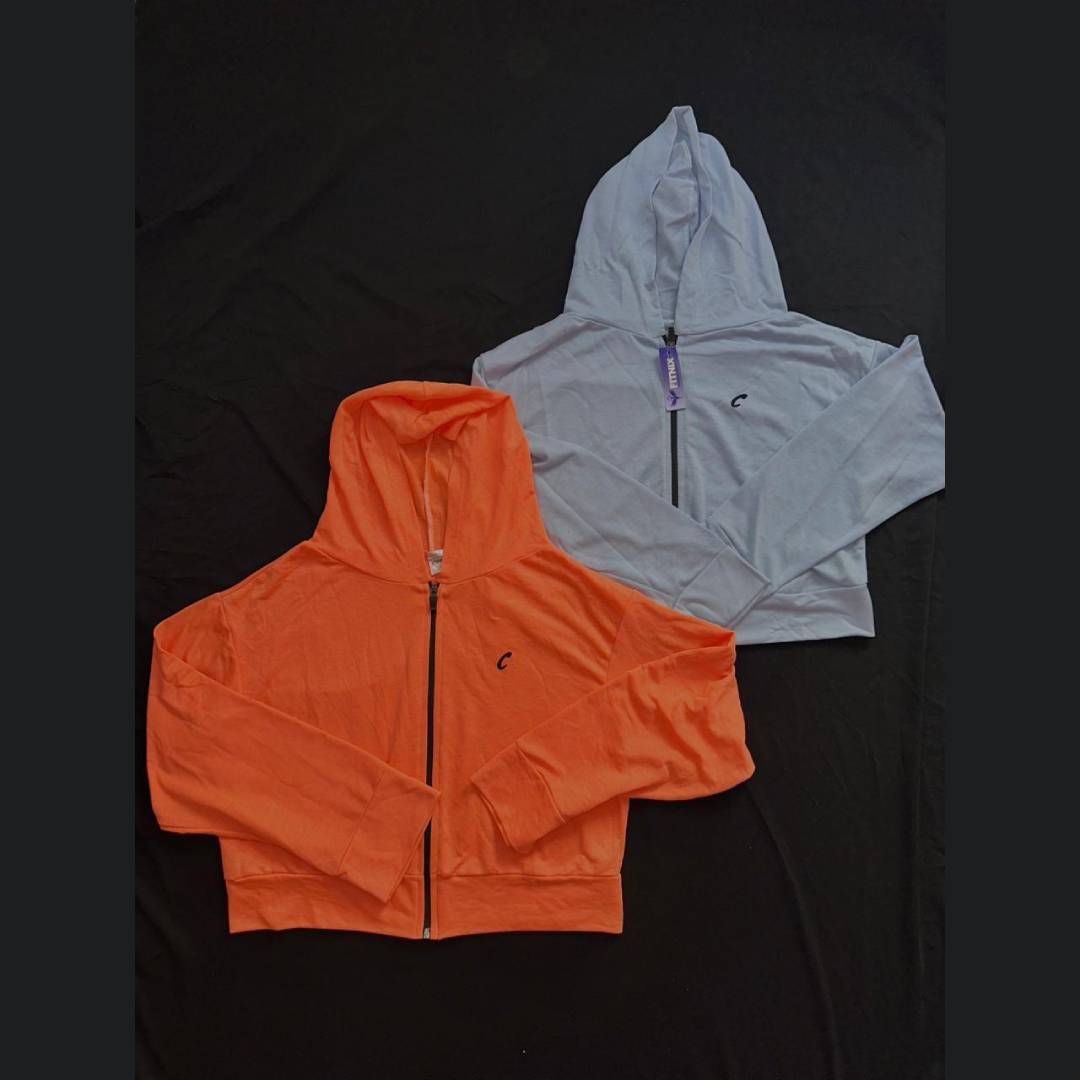 Dos sudaderas cortas con cremallera, una naranja y otra azul claro, sobre tela negra.