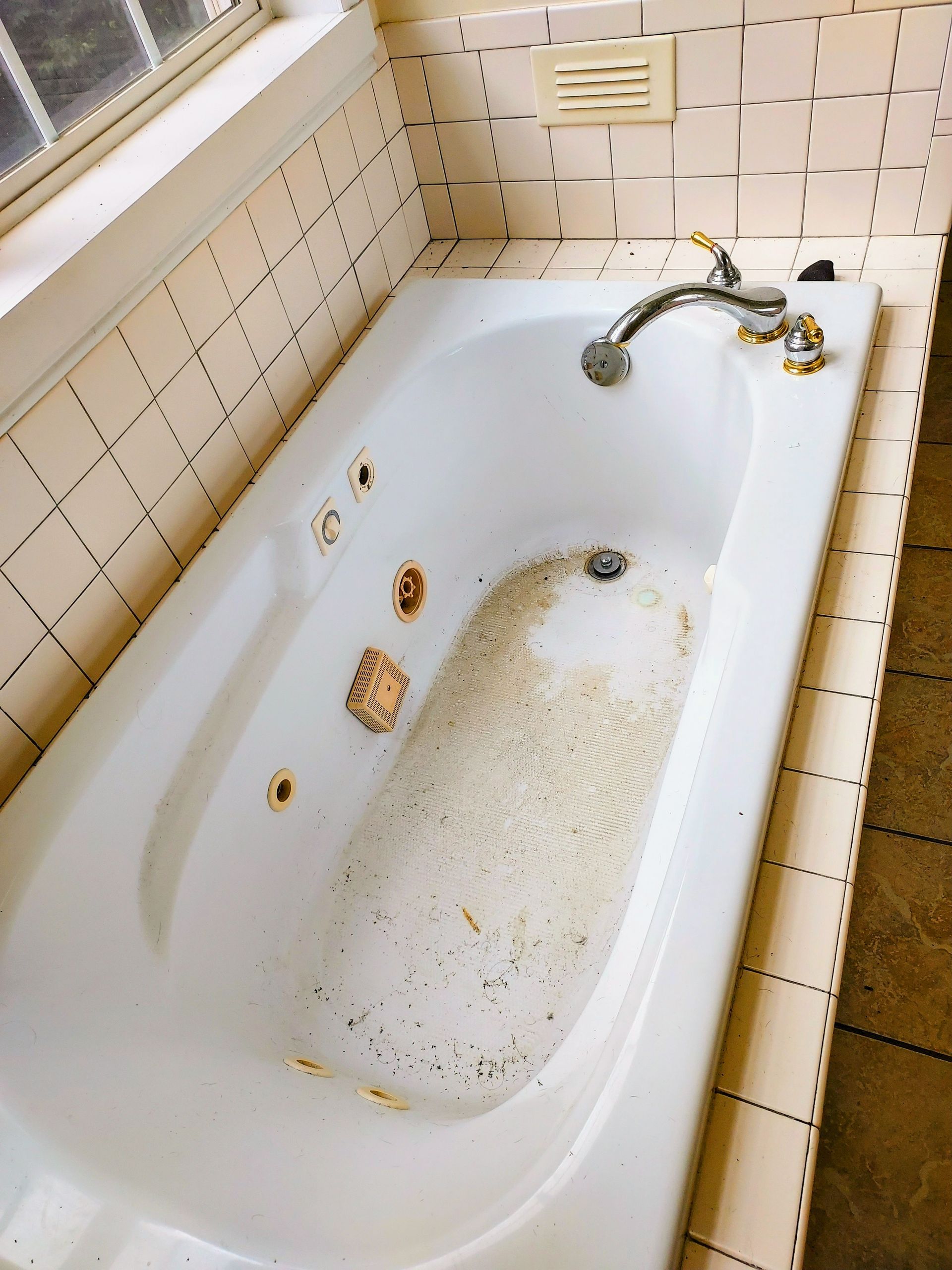 White jacuzzi tub with dirt and debris, against beige tiled walls.