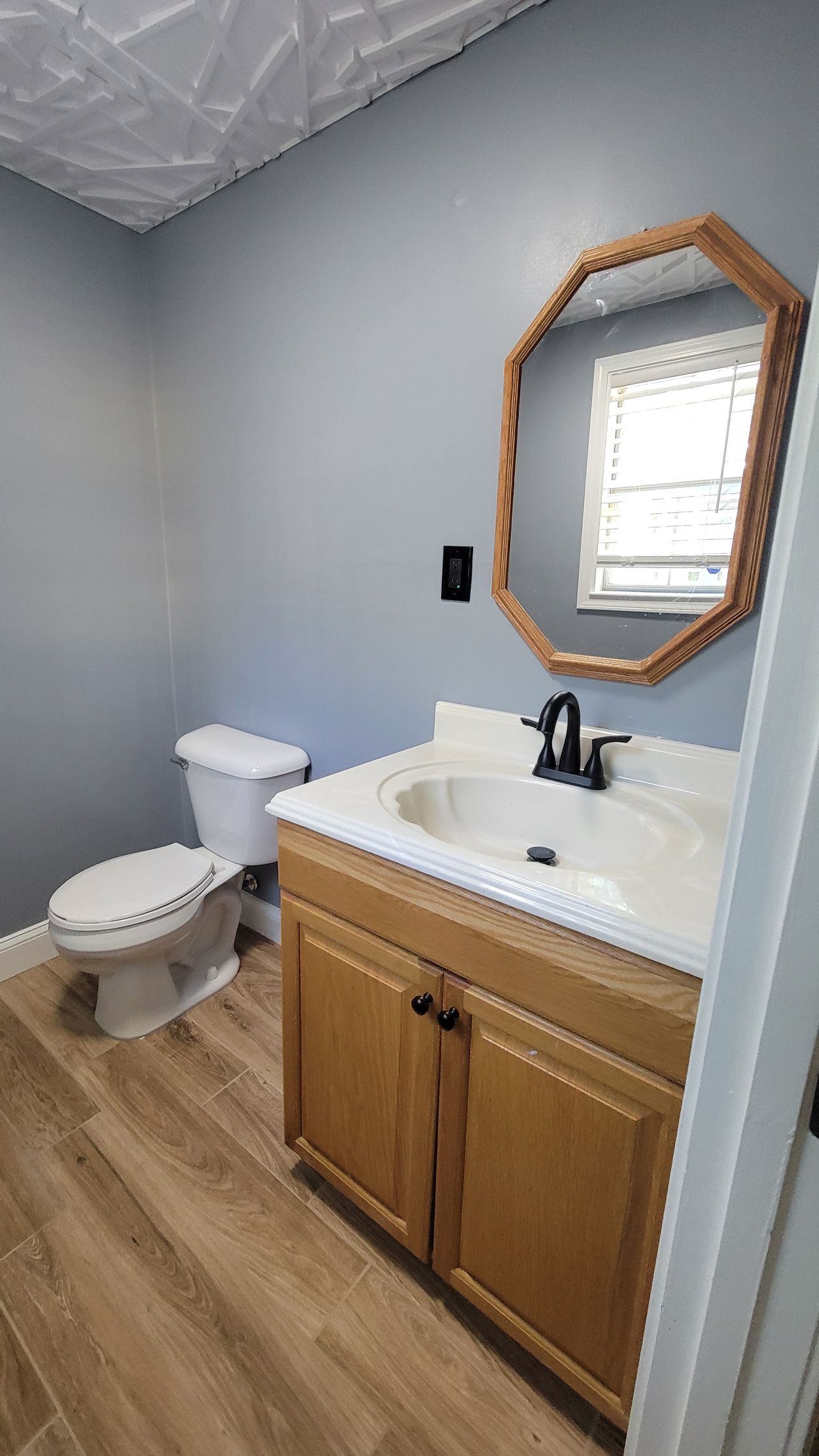 Bathroom with light blue walls, wooden vanity, octagonal mirror, toilet, and light wood flooring.