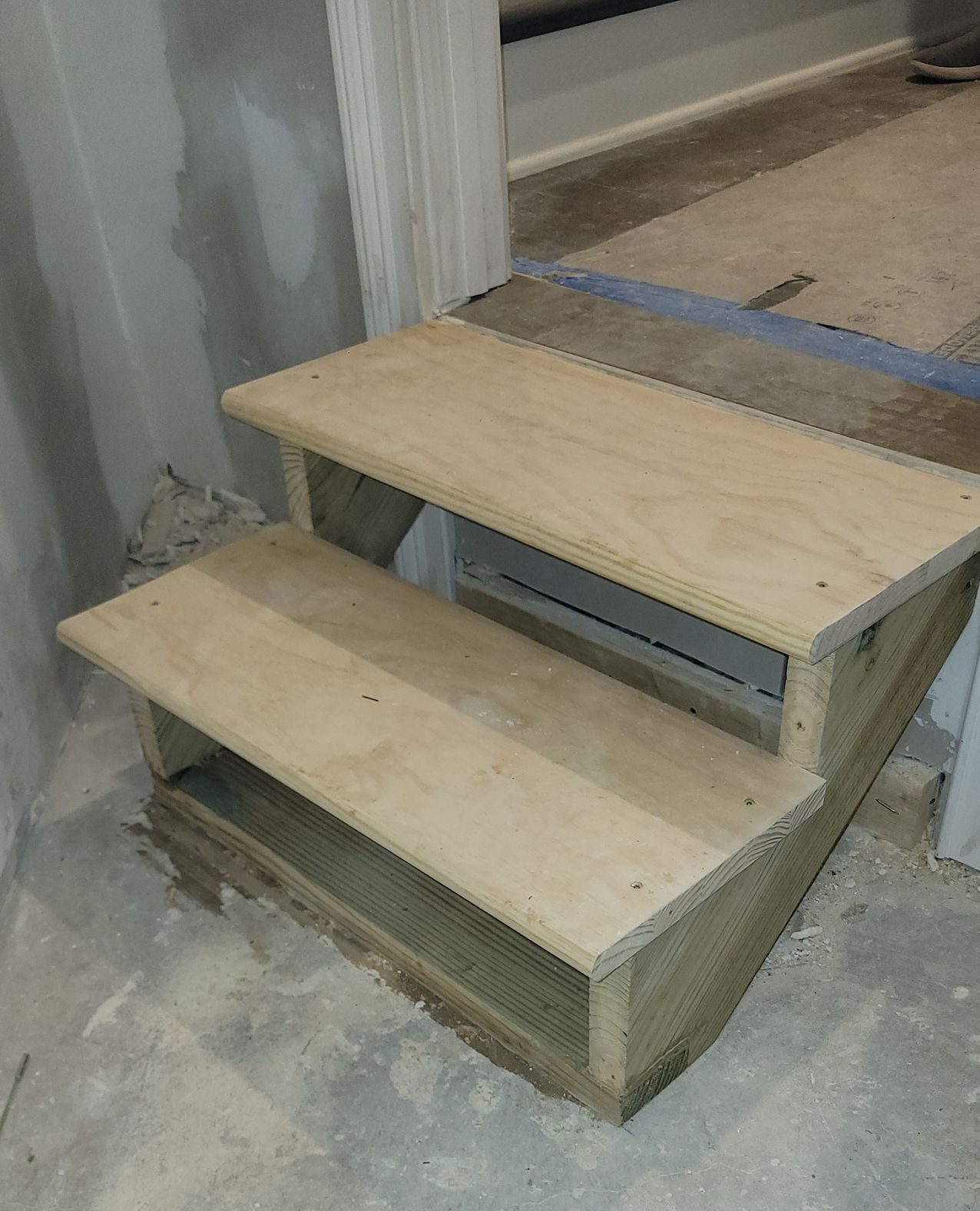 Two-step wooden staircase under construction in a room with concrete flooring and walls.