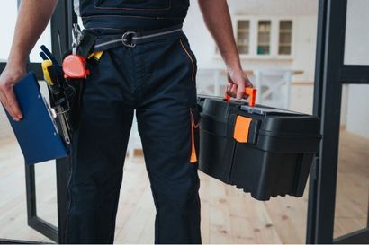 Person in blue overalls holding a toolbox and clipboard, entering a building.