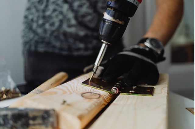 Person uses a drill to screw a hinge onto a wooden board.