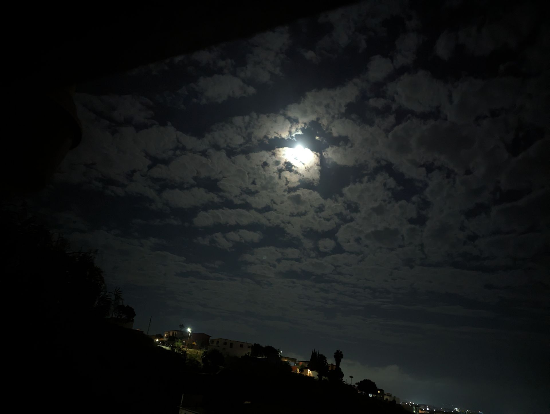 Night sky with full moon peeking through clouds, illuminating the dark landscape of a town.
