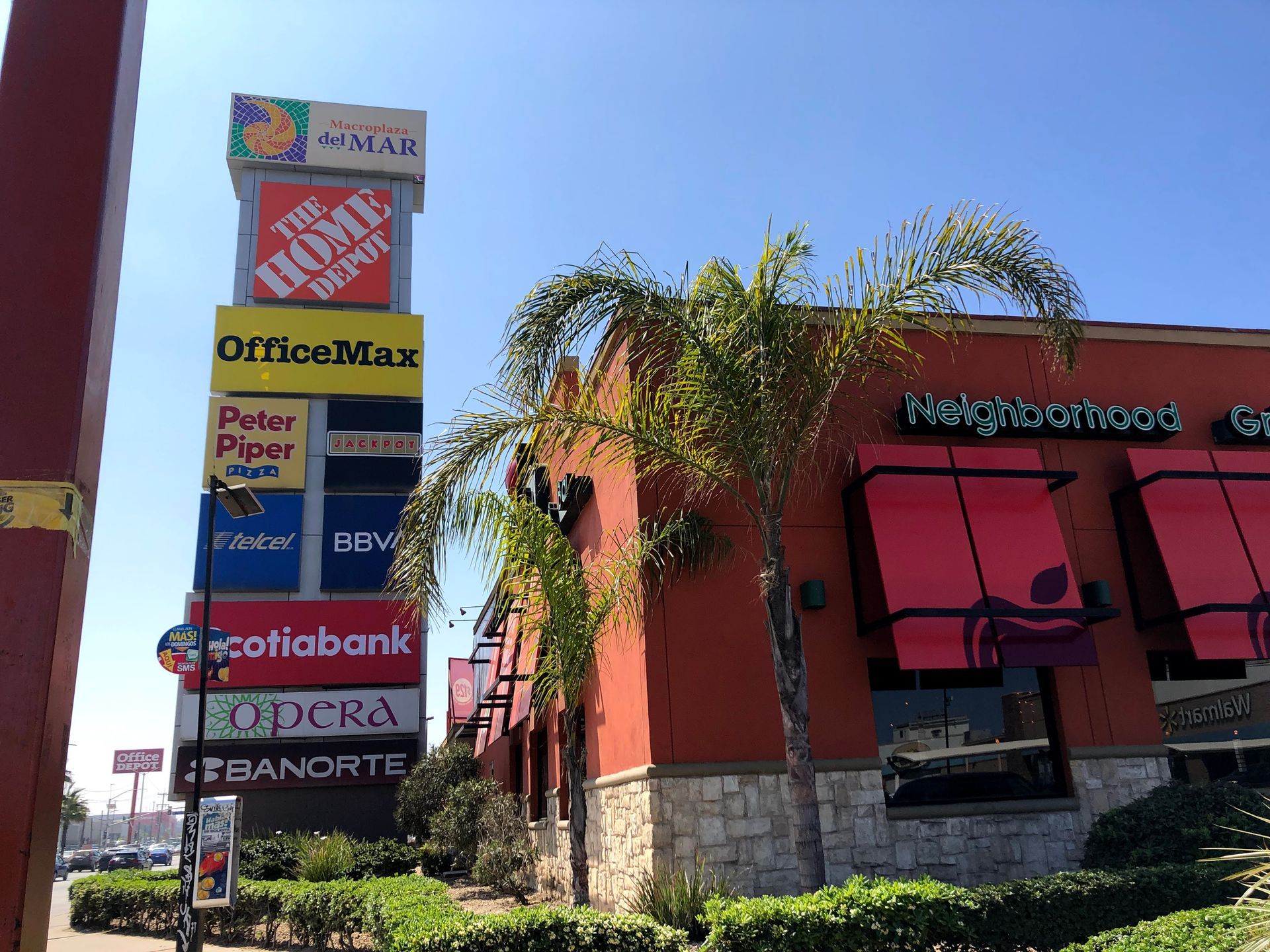 Sign tower with store logos next to a red stucco restaurant with awnings and palm trees.