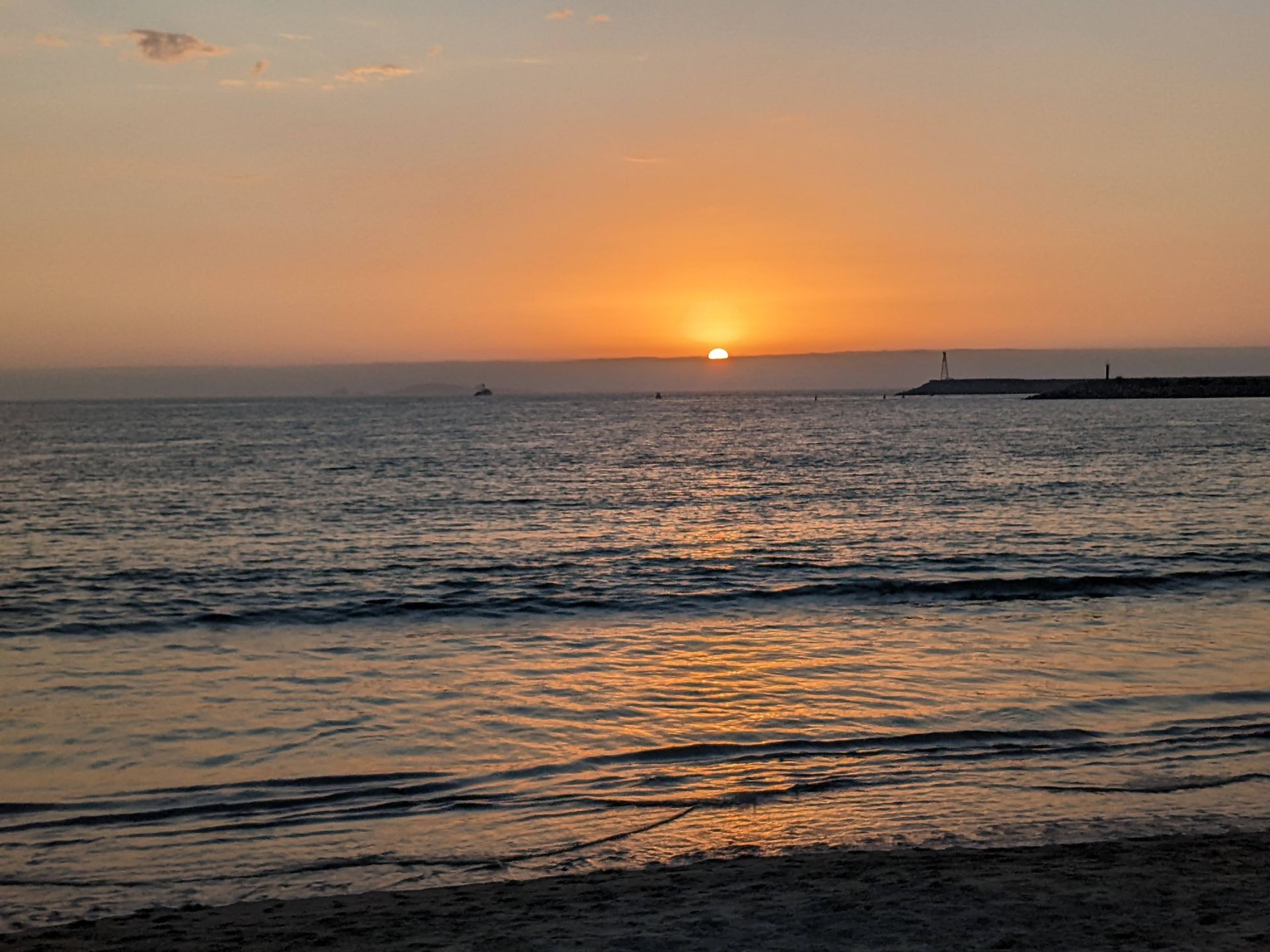 Sunset over the ocean, orange sky reflecting on water, waves on shore, silhouette of land and boat.