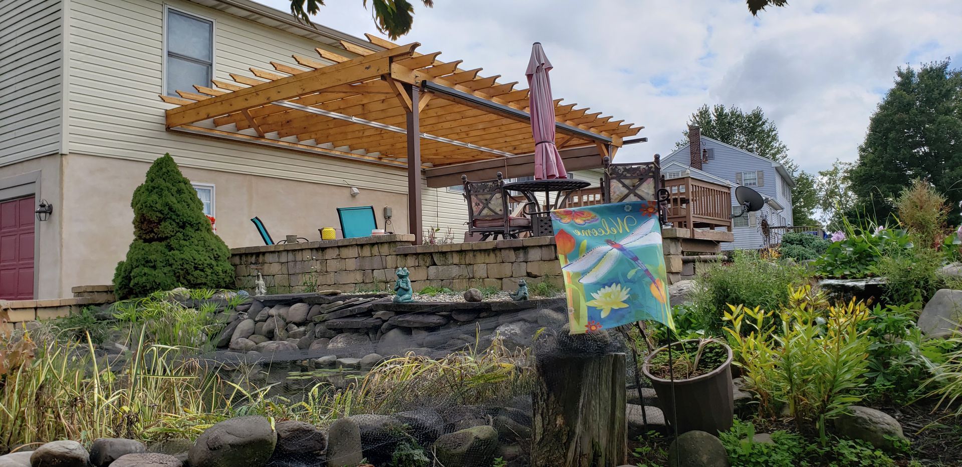 Outdoor patio with wooden pergola, stone steps, and colorful sign in a garden beside a house
