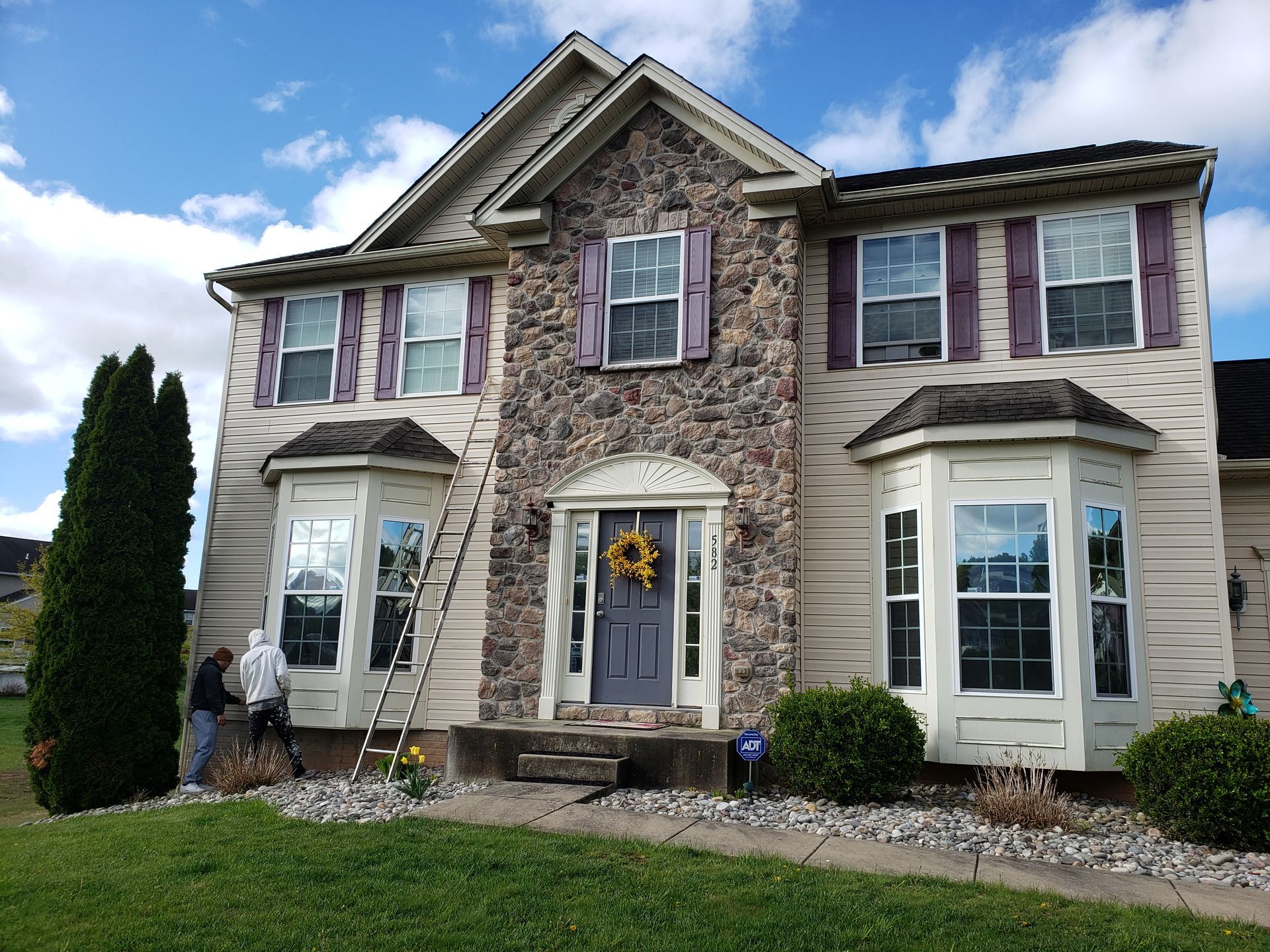 Two-story beige house with purple shutters, stone facade, and a front lawn under a blue sky