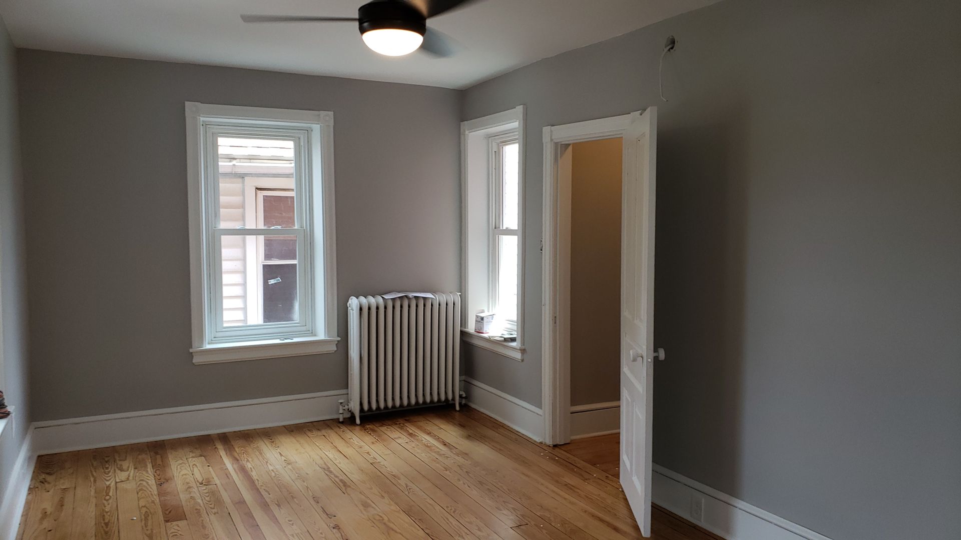 Empty gray room with hardwood floors, two windows, radiator, and an open door.