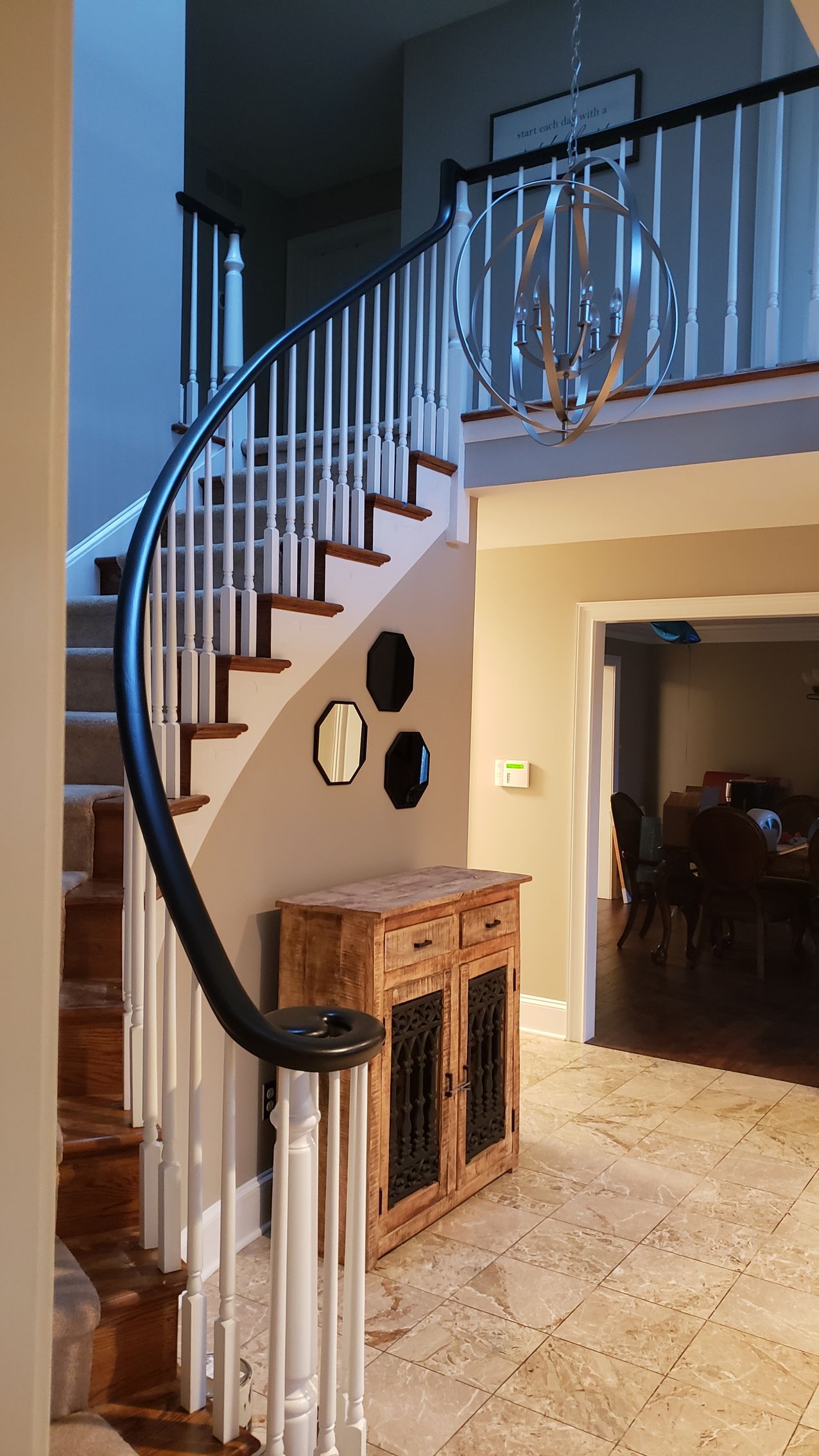 Curved staircase with dark railing in a warmly lit home entryway, beside a small wooden cabinet.