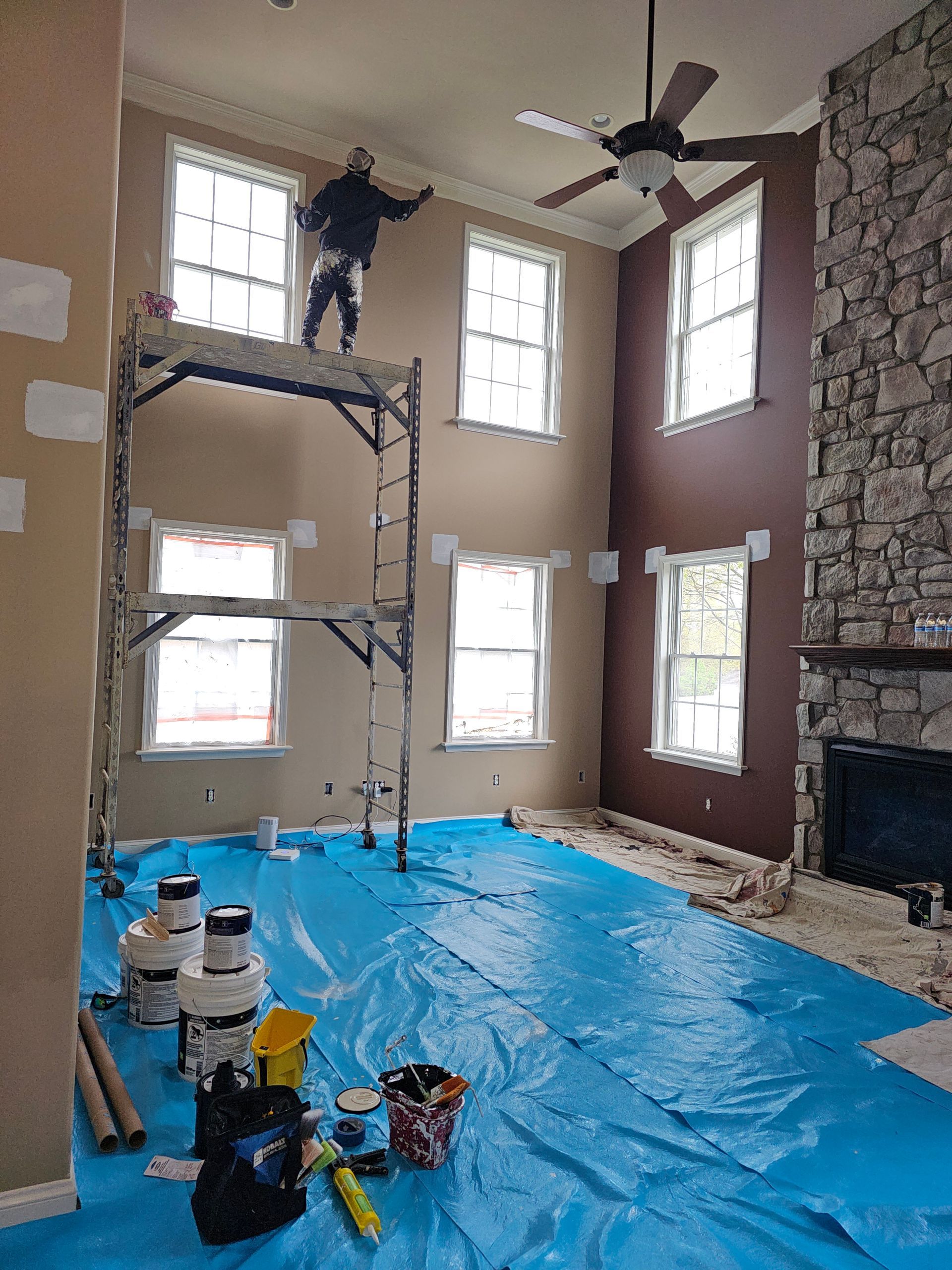 Worker on scaffolding inside a tall room, with blue tarp floor covering and stone fireplace wall.