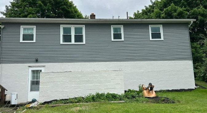 Two-story gray house with white lower siding, four windows, and a grassy yard with a small tree stump.