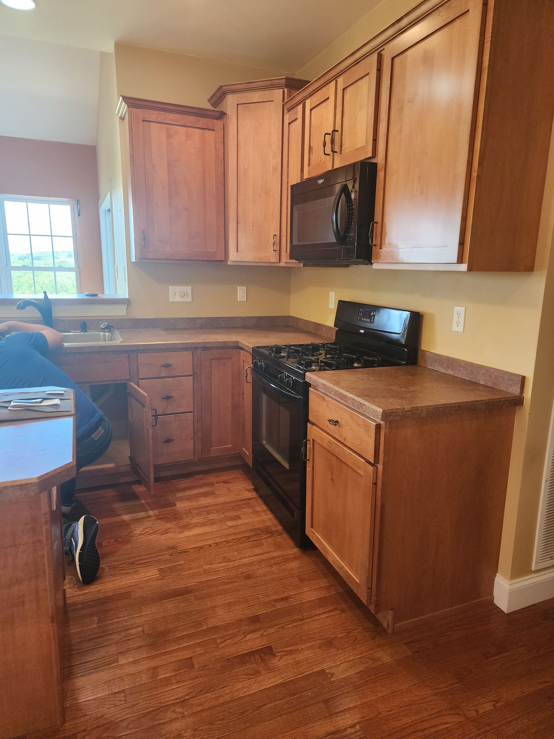Wooden kitchen with cabinets, black stove and microwave, and a window by the sink.