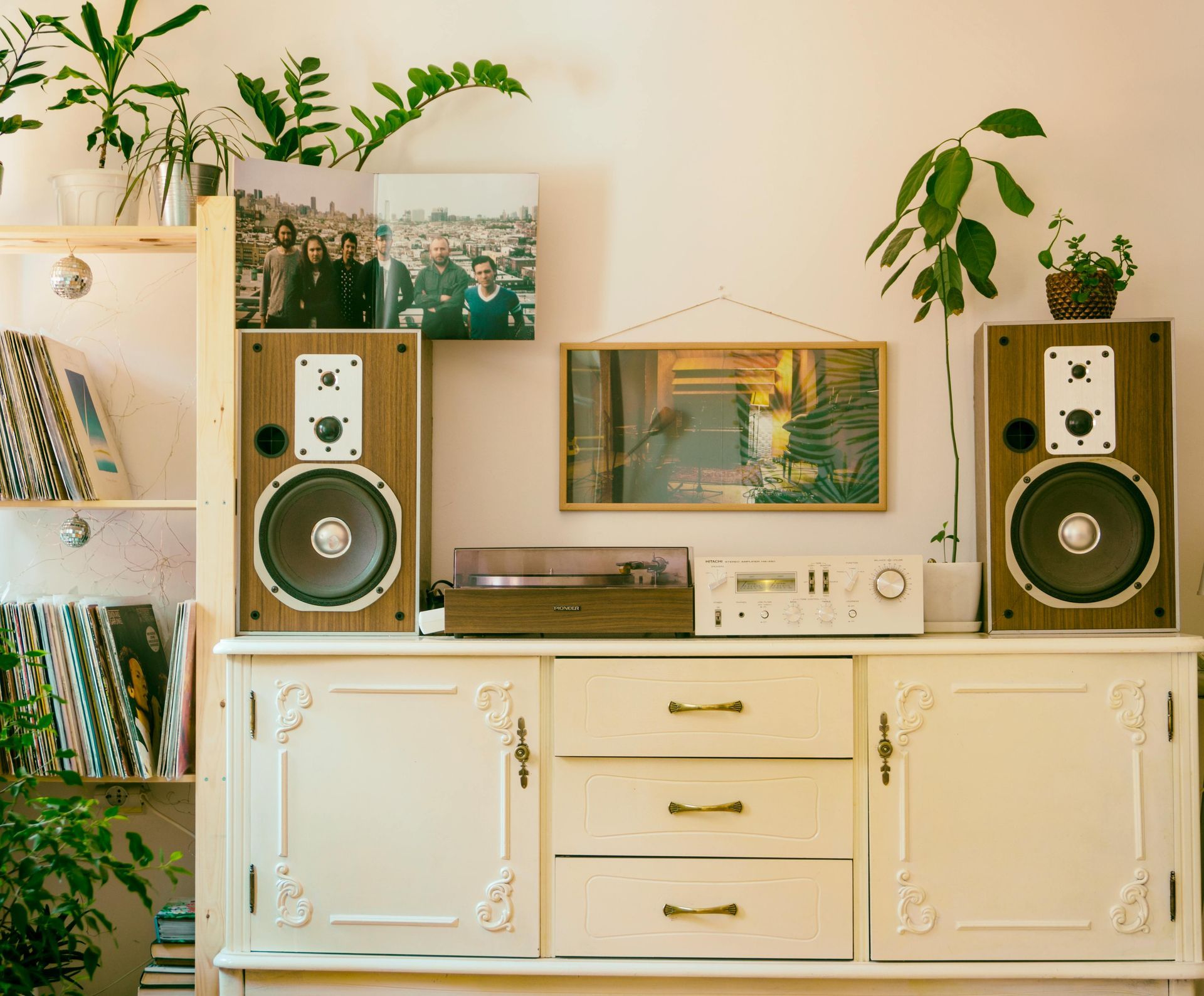 Vintage stereo setup on white cabinet with plants, framed art, and two large speakers in a cozy room