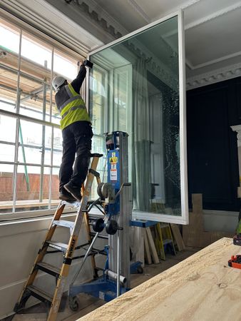 A worker in a high-visibility vest uses a mechanical lift to install a large window pane in a room with ornate molding.