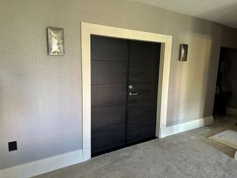 Double dark wood doors with a white frame and moldings on a light gray wall with two wall sconces and carpeted floor.