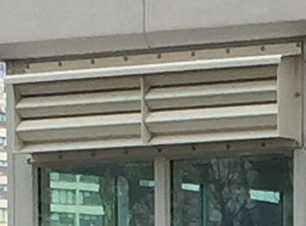 A beige, horizontal metal ventilation louver mounted above a glass window on a building exterior.