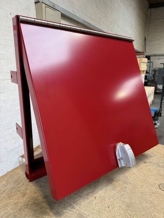 A red, hinged metal access door or hatch with a silver latch, shown sitting on a workbench in an industrial setting.