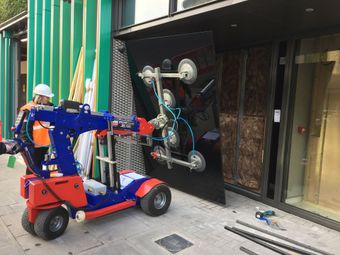 A worker operates a blue and red robotic glass lifter to install a large, black glass panel into a building facade.