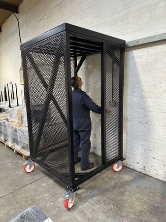 A person in a dark jumpsuit stands inside a tall, black metal cage on wheels in a warehouse setting.