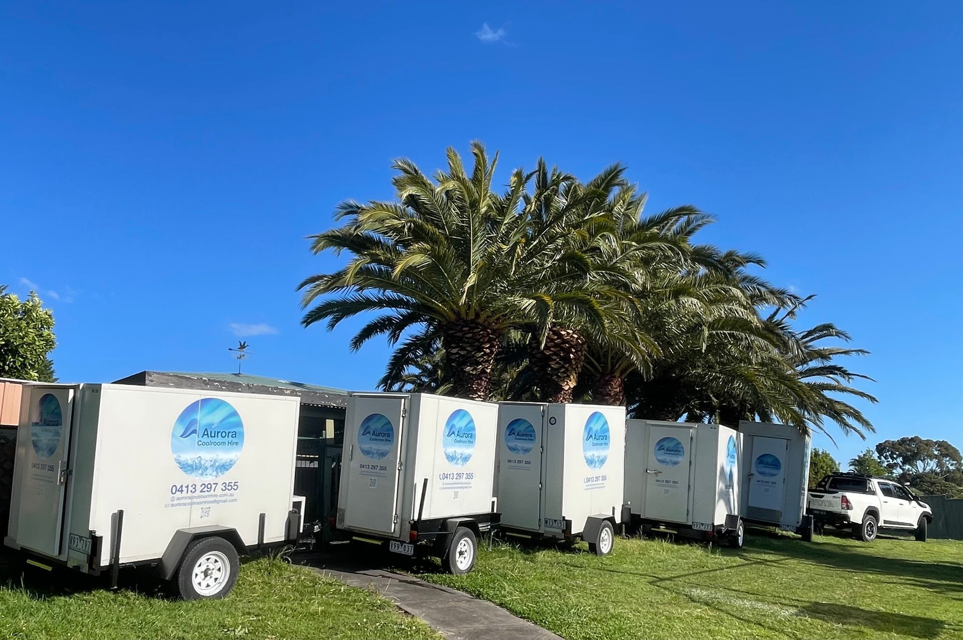 White trailers with blue logos parked on grass, palm trees in background, blue sky.
