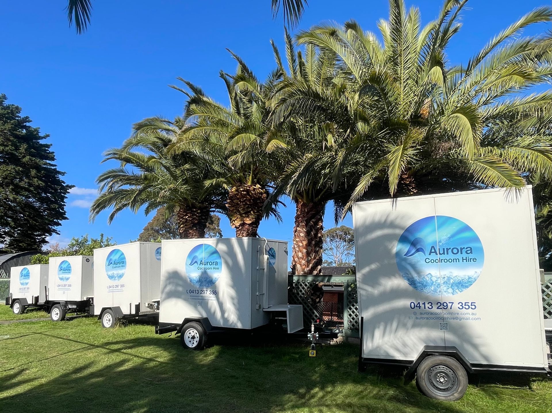 Line of white portable coolrooms parked on grass, with palm trees in the background. Each has a logo.