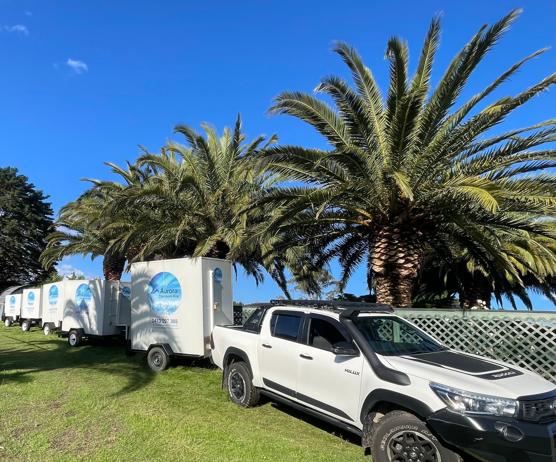 A white truck towing a line of small, white trailers under palm trees on a grassy field, against a blue sky.