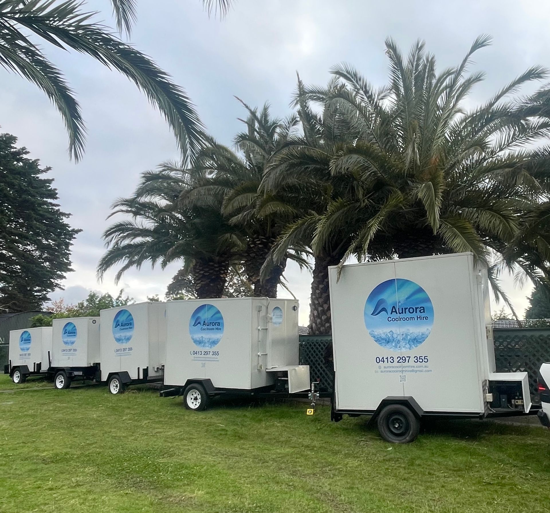 Five white portable coolroom trailers parked on grass, palm trees in the background.