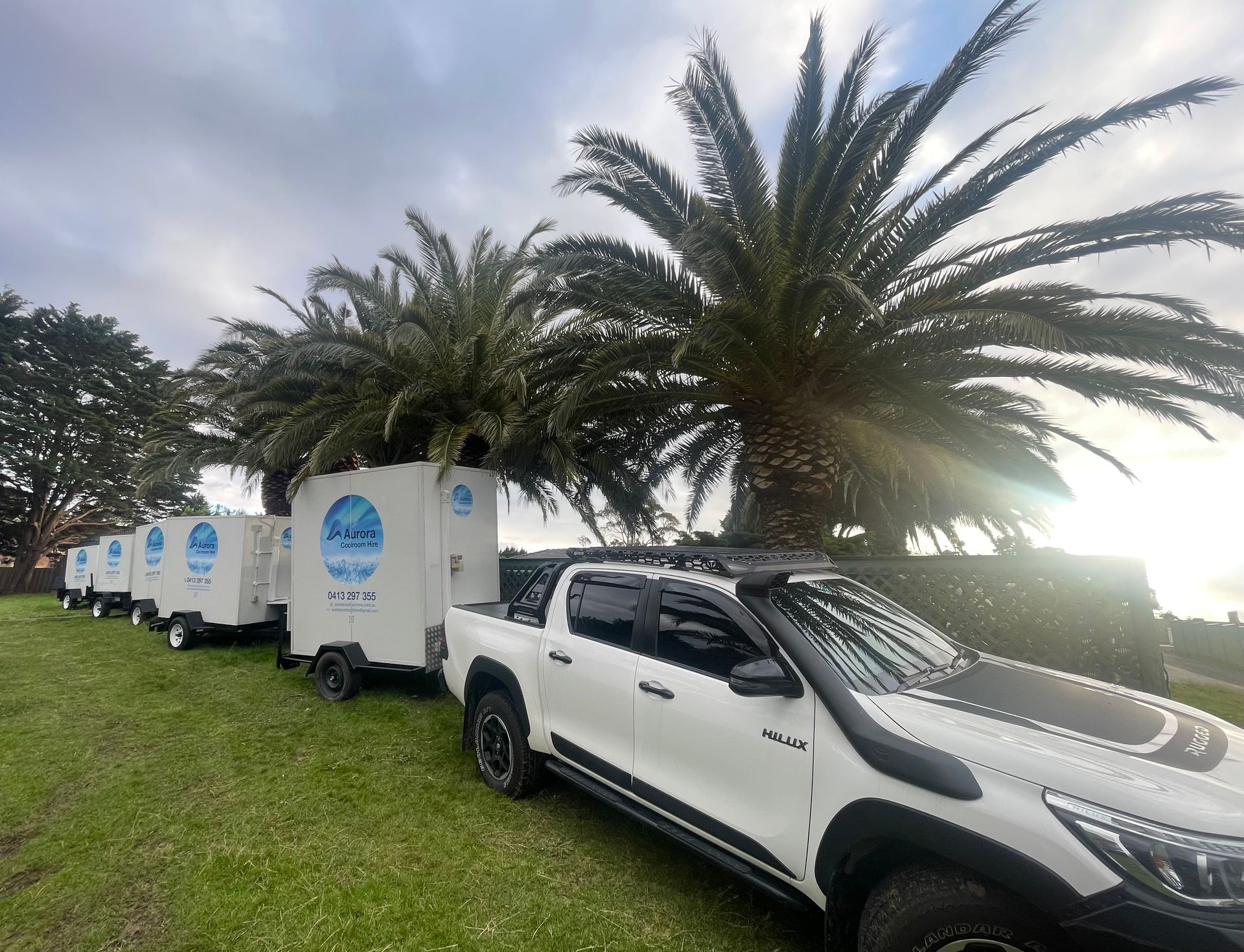 A white pickup truck towing a line of white trailers on grass, under palm trees.