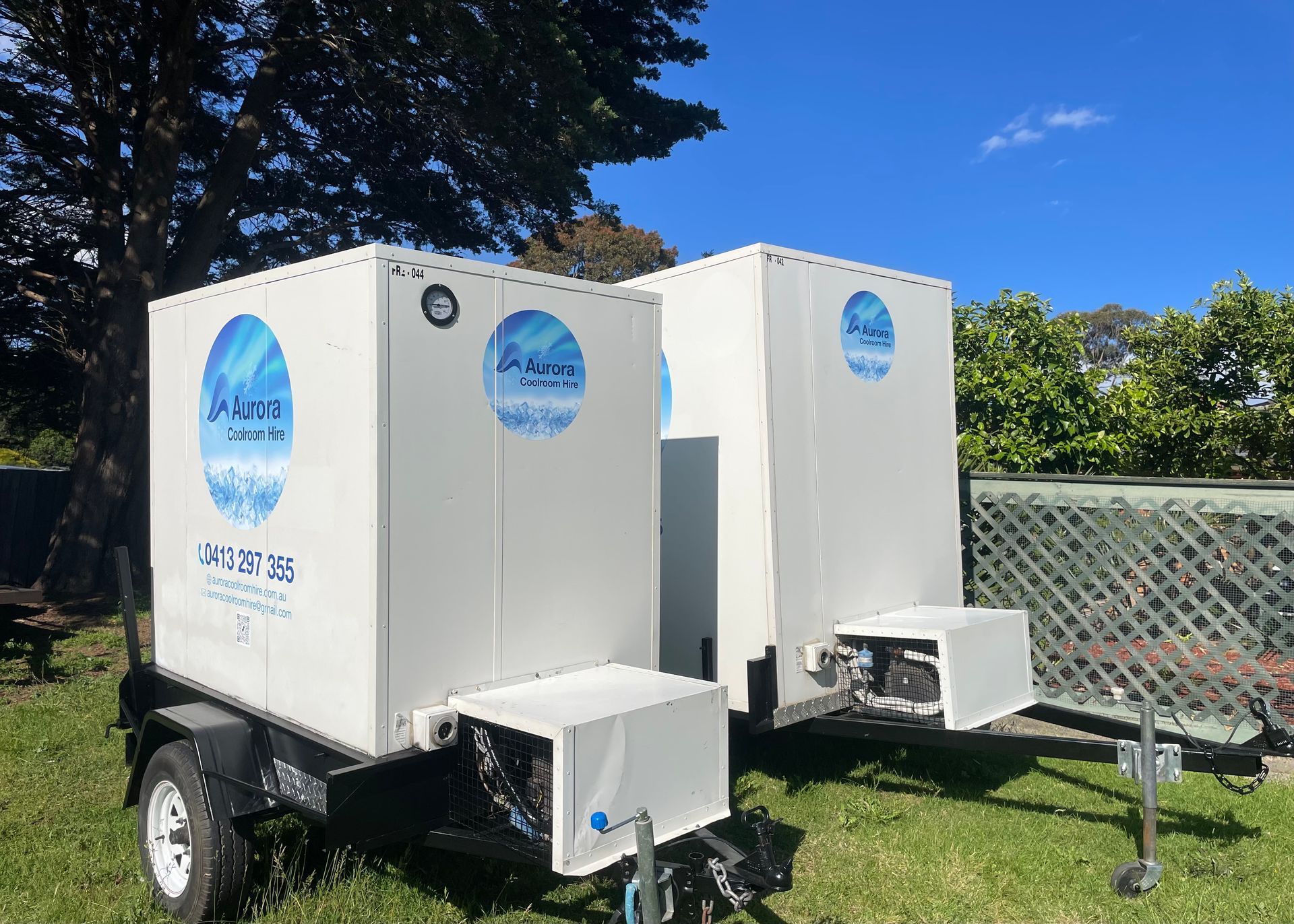Two white rectangular coolroom machines on trailers, parked on grass.