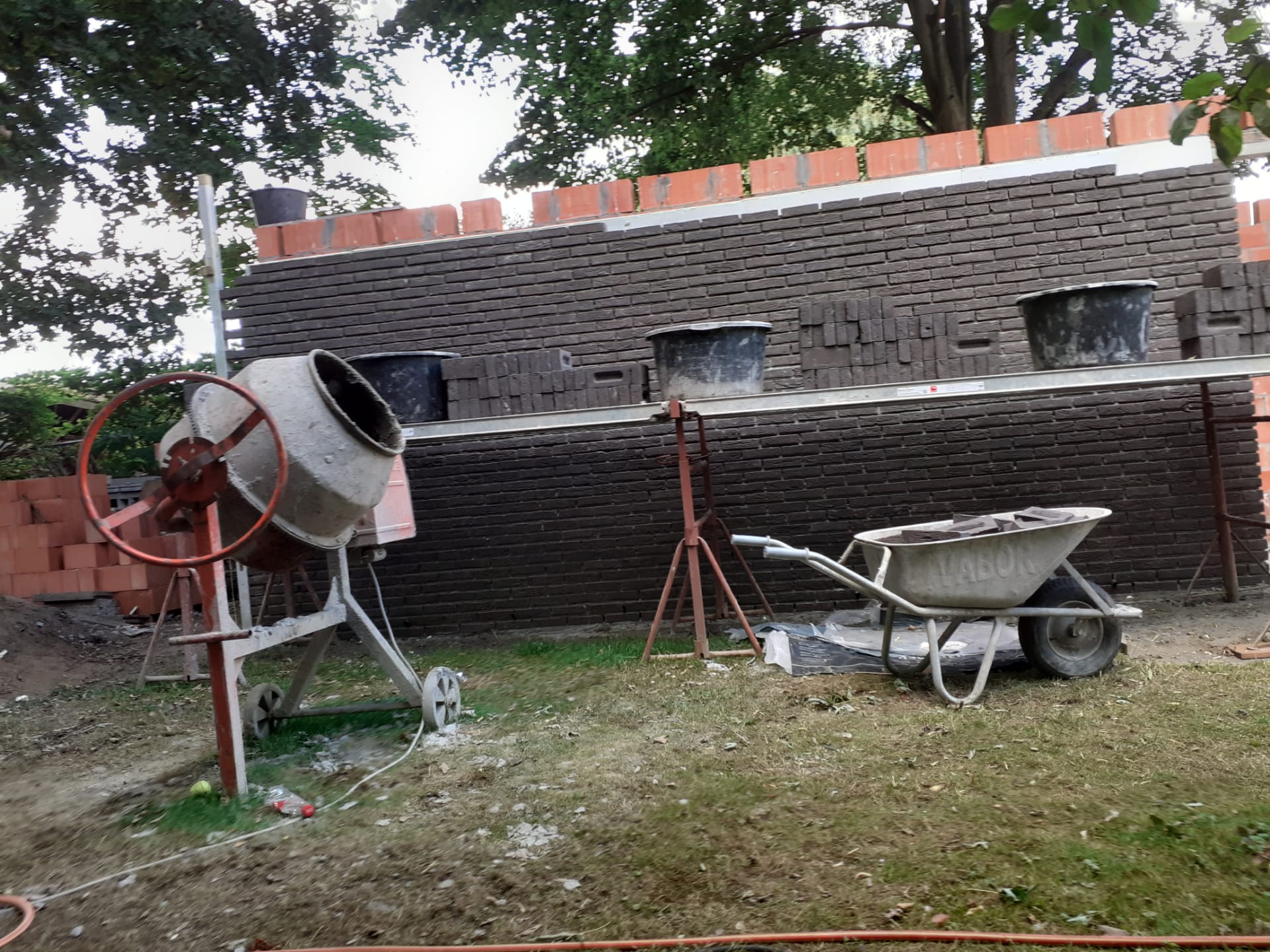 Voor een bakstenen muur staan ​​een cementmixer en een kruiwagen.