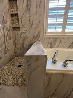 Bathroom with marble-patterned walls, a shower, and a bathtub near a window with closed blinds.