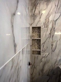 Marble-tiled shower with built-in shelf. White and gray veining. Glass shower door and black shower fixtures.