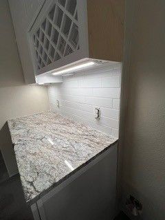 Kitchen counter with granite countertop, white tile backsplash, and overhead lighting. Wine rack above.