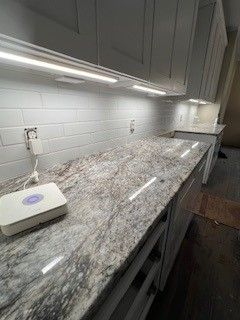 Kitchen with grey cabinets, white tile backsplash, and granite countertops, lit by under-cabinet lights.