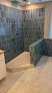 A walk-in shower with gray-blue tiled walls, a tan step, and tan floor tiles, next to a light-colored vanity.
