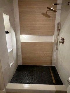 Shower with pebble floor, wood-look tiled wall, built-in shelf, and gold-toned shower head.