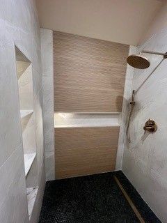 Shower with textured brown tile wall, shower head, shelves on the left, and dark floor.