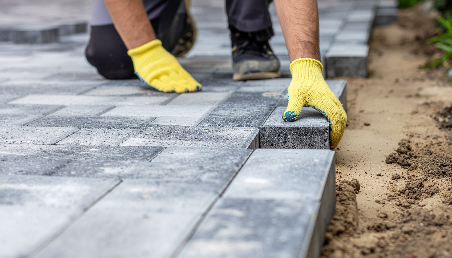 A person wearing yellow gloves placing a grey paver to build a walkway.
