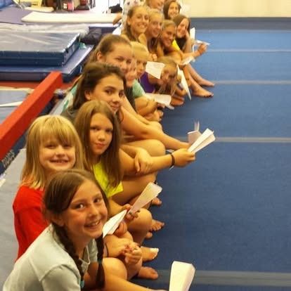 Children sitting in a row, smiling and holding paper airplanes on a blue mat.
