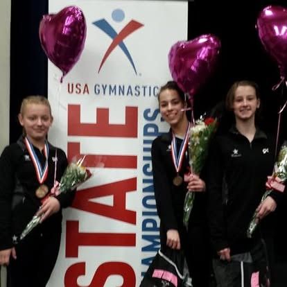 Gymnasts at a USA Gymnastics State Championship, smiling, holding bouquets and medals. Two pink heart balloons in background.