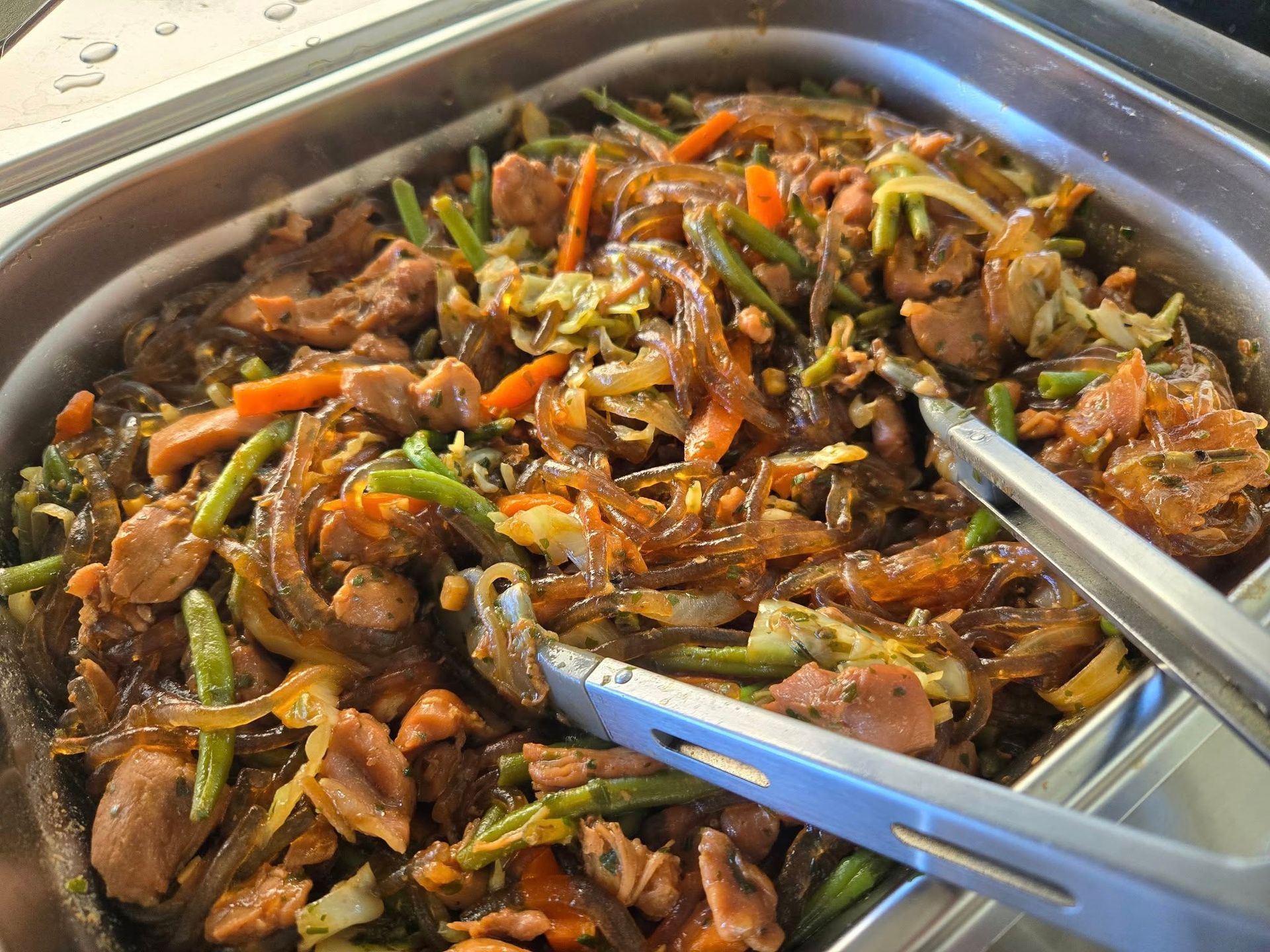 A metal buffet pan filled with stir-fried glass noodles, sliced chicken, green beans, carrots, and onions with metal tongs.