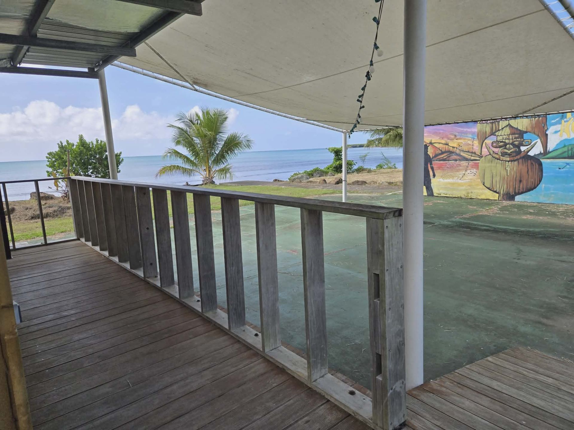 A wooden deck with a low railing overlooks a calm sea, featuring a shaded patio area with a mural in the background.