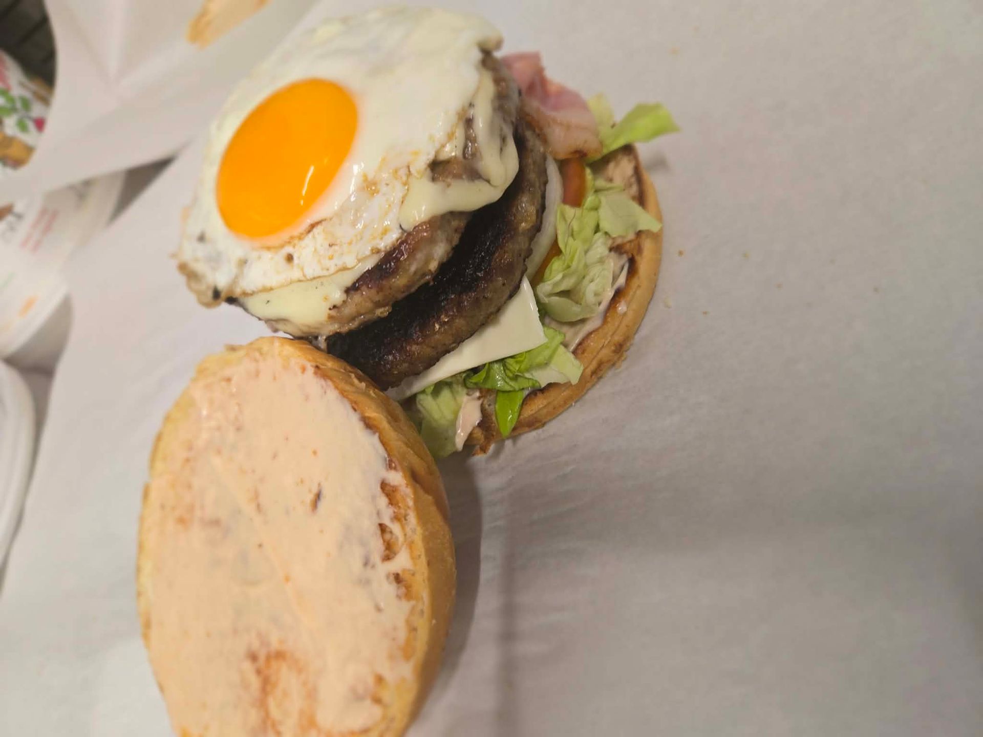 An open double cheeseburger topped with a fried egg, lettuce, and a smear of pink sauce on the bun, served on white paper.
