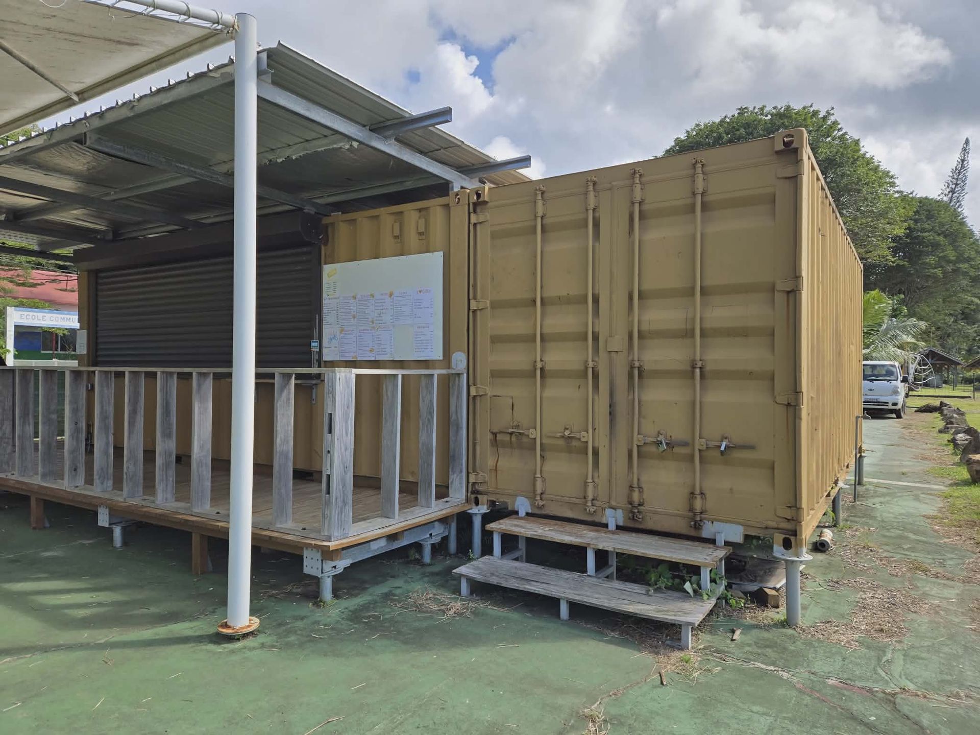 Tan shipping container building with a covered wooden porch, rolling shutter door, and steps, set on a green paved lot.