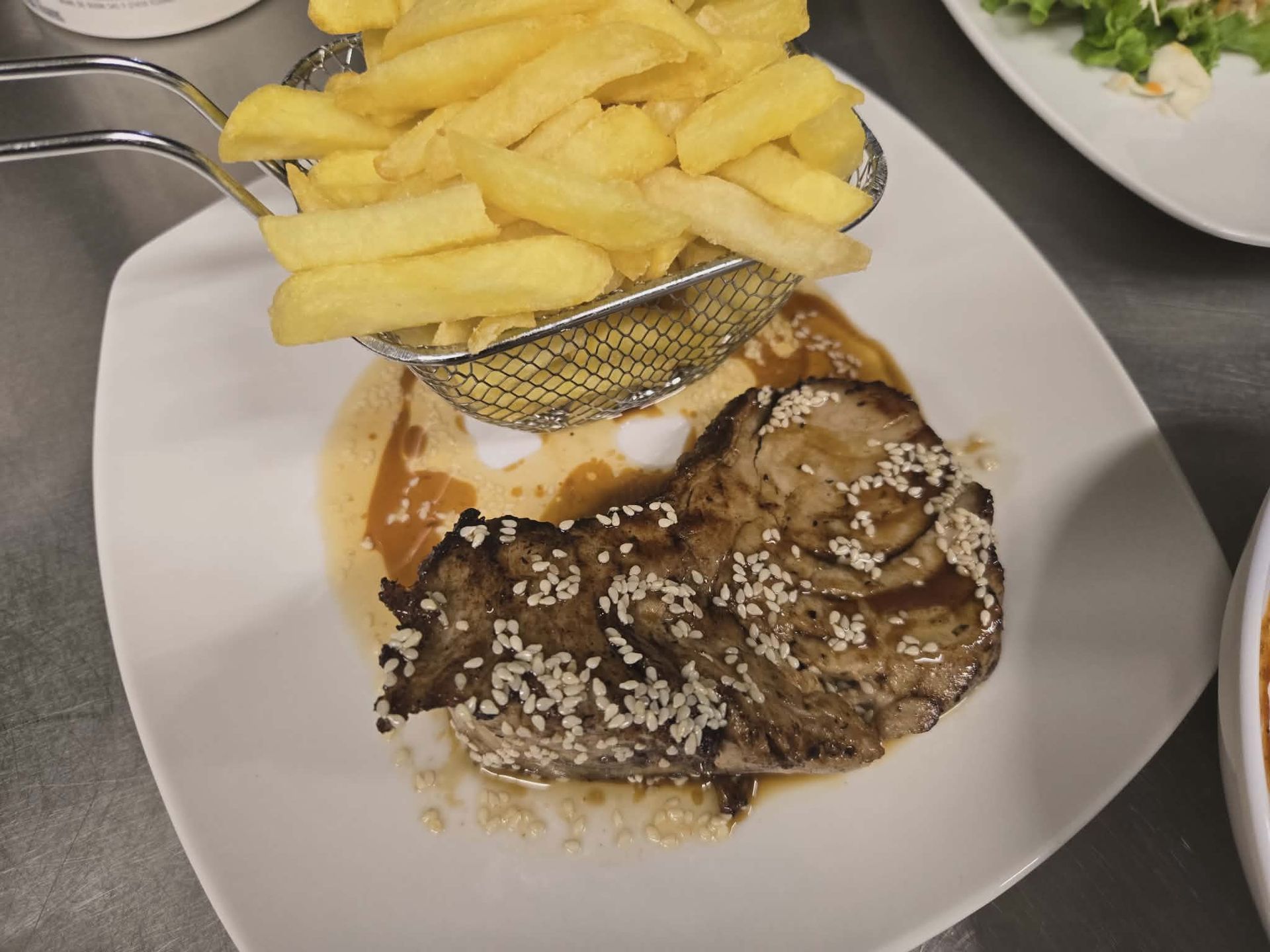 A grilled steak topped with sesame seeds served on a white plate with a metal basket of French fries.