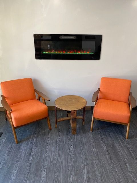 Two orange armchairs and a small round table face a fireplace on a white wall.