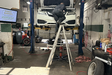 Mechanic on a ladder working on a white car raised on a lift inside a garage.