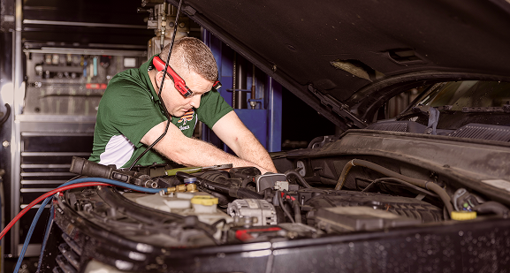 Mechanic working on a car engine, wearing safety glasses and a green shirt.