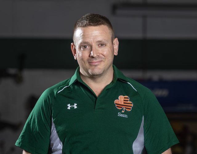 Man in green polo shirt, three-leaf clover logo, smiling slightly, indoor setting.