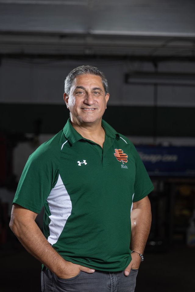 Man in green polo shirt, hands in pockets, smiling indoors.